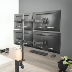 Four black monitors mounted on a desk stand in a modern office setup with keyboard and plants.